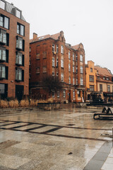 A rainy day view of a modern urban square with a wet pavement reflecting light, framed by a mix of new and old brick buildings. The image conveys a moody, atmospheric urban scene.