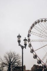  An antique three-headed street lamp stands tall beside a large Ferris wheel, with bare trees and historic buildings in the background under a plain, overcast sky.