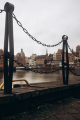A low-angle shot of a weathered wooden dock with a rustic chain barrier, overlooking a blurred European cityscape and river on a cloudy day.