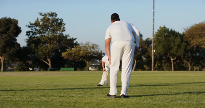 Diverse male cricket players under-arming, fielding and lobbing ball past stumps in park practice