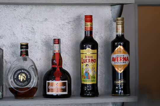 Italian liqueur collection featuring Vecchia Romagna brandy, Grand Marnier, Amaro Lucano, and Averna, lined up on wooden shelf with muted gray backdrop