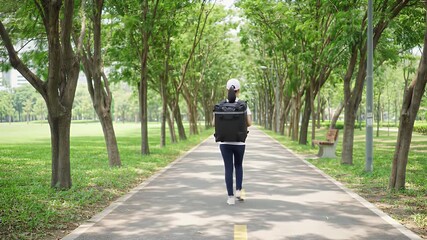 Back View of a Young Asian Delivery Courier in White Uniform and Black Backpack Walking Down a Tree-Lined Path on a Sunny Afternoon Perfect for Takeaway Order Concepts and Delivery Advertisement