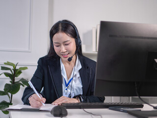 An Asian woman in a suit and headset is taking notes at a computer. Look like a call center