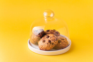 Stack of fresh chocolate American style chip cookies with nuts and chocolate drops on a tray with transparent lid on yellow background. Cookies with peanut butter filling inside a glass jar