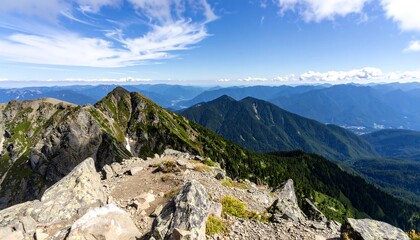 Panoramic mountain vista; rocky foreground, lush green slopes, hazy distant ranges under a bright blue sky