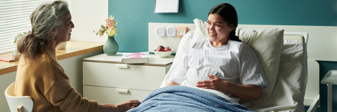 Header of young pregnant Caucasian woman lying in hospital bed smiling and talking with senior mother sitting nearby, both interacting in medical setting with visible medical equipment - Powered by Adobe