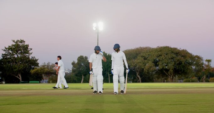 Completing run, diverse male cricketers high-fiving and walking off dusk pitch carrying bats