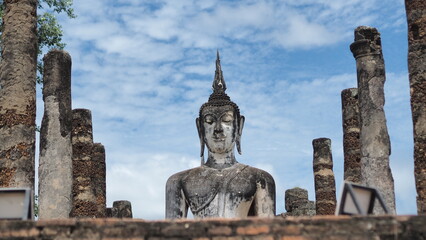 Obraz premium A low-angle, medium shot of a large, weathered, standing Buddha statue. The statue is enclosed within the crumbling brick and light-colored stone walls of a temple ruin. The statue's face is weathered