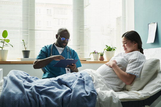 Black middle aged male doctor writing on clipboard while sitting beside pregnant Caucasian young woman lying in hospital bed, both interacting during prenatal checkup
