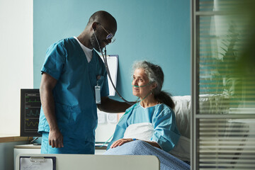 Obraz na płótnie Canvas Black male young doctor examining senior Caucasian woman patient with oxygen tube using stethoscope in hospital room, medical monitor displaying vital signs nearby