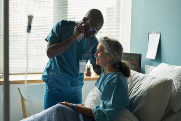 Black male middle aged doctor examining senior Caucasian woman patient with oxygen tube in hospital bed, doctor listening to patient chest and lungs with stethoscope, medical monitor in background