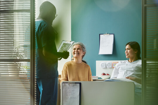 Middle aged Caucasian woman smiling while sitting on hospital bed next to young pregnant Hispanic woman, Black male nurse standing nearby holding clipboard and talking during visit at maternity clinic