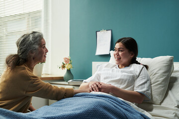 Middle aged Caucasian mother sitting beside hospital bed holding hand of young pregnant woman lying under blanket smiling, comforting daughter before childbirth and making eye contact in patient room