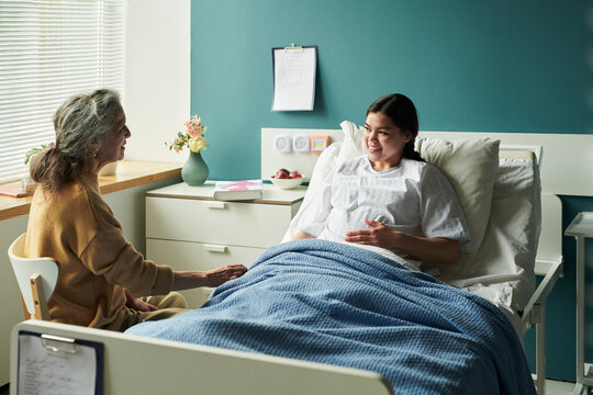 Young Caucasian woman lying in hospital bed, smiling and touching belly, while middle aged mother sitting beside bed holding her hand in supportive gesture during visit