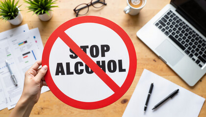 Hand holding stop alcohol sign on wooden table with laptop and plants  