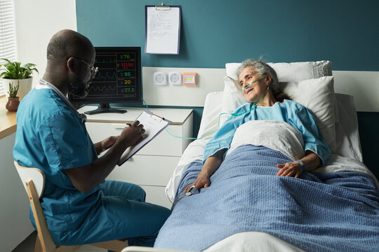 Senior Caucasian woman lying in geriatric hospital bed receiving oxygen therapy while Black male nurse sitting nearby writing notes on clipboard, medical monitor displaying vital signs - Powered by Adobe