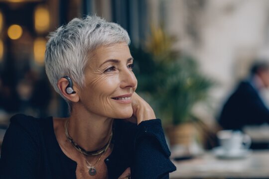 Stylish mature woman enjoying coffee with wireless earbuds in cafe - Powered by Adobe