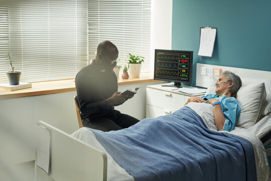 Black middle aged male priest sitting beside hospital bed talking to senior Caucasian woman lying under blanket with medical monitor in background, reading Bible, both engaging in faith conversation - Powered by Adobe