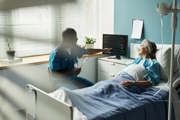 Middle aged Black man doctor explaining vital signs on monitor to senior Caucasian woman patient lying in hospital bed with oxygen tube, medical equipment visible in background