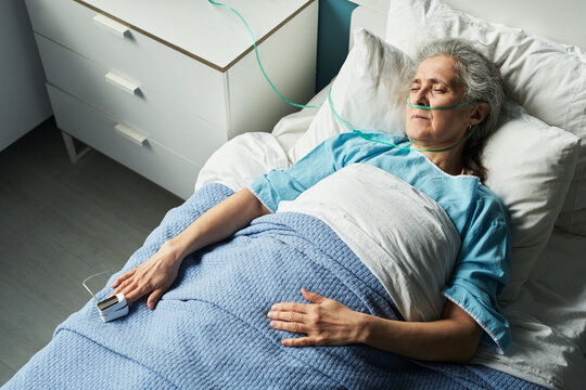 Senior Caucasian woman lying in geriatric hospital bed with oxygen tube and monitoring device on finger, resting with eyes closed, showing signs of illness or recovery in medical setting - Powered by Adobe