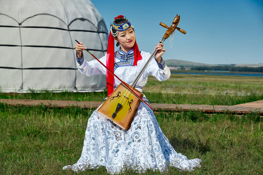 A Mongolian female musician in traditional Mongolian attire playing the morin khuur (horsehead fiddle) in front of a yurt.