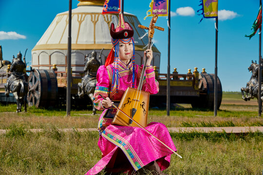 A Mongolian female musician in traditional Mongolian attire playing the morin khuur (horsehead fiddle) in front of a yurt.