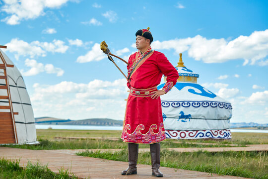 Mongolian men dressed in traditional Mongolian attire and holding bows and arrows stand in front of a yurt.