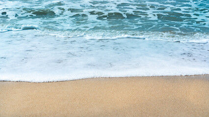 a closeup beach detail reveals the texture of the golden sand as gentle turquoise waves with white foam wash over the shoreline.