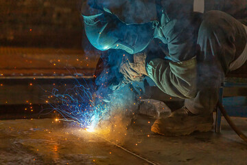 A male welder wearing a fireproof and protective welding mask welds a huge seam in a sheet of metal
