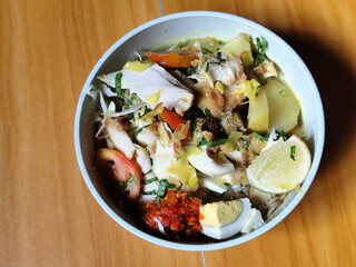 A bowl of Indonesian chicken soto with shredded chicken, sliced boiled egg, vermicelli, sliced cabbage, bean sprouts, celery, chili sauce and lime, mixed with delicious sauce. On a wooden table.	
