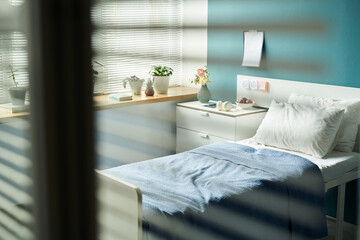 Hospital room featuring unoccupied single bed with blue blanket, bedside table holding flowers and medical equipment, window with blinds partially open, potted plants on windowsill