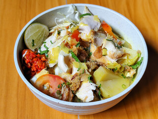 A bowl of Indonesian chicken soto with shredded chicken, sliced boiled egg, vermicelli, sliced cabbage, bean sprouts, celery, chili sauce and lime, mixed with delicious sauce. On a wooden table.	

