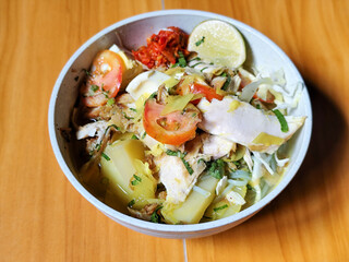 A bowl of Indonesian chicken soto with shredded chicken, sliced boiled egg, vermicelli, sliced cabbage, bean sprouts, celery, chili sauce and lime, mixed with delicious sauce. On a wooden table.	
