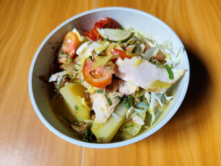 A bowl of Indonesian chicken soto with shredded chicken, sliced boiled egg, vermicelli, sliced cabbage, bean sprouts, celery, chili sauce and lime, mixed with delicious sauce. On a wooden table.	
