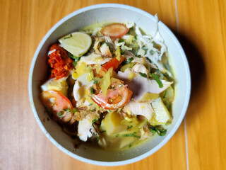 A bowl of Indonesian chicken soto with shredded chicken, sliced boiled egg, vermicelli, sliced cabbage, bean sprouts, celery, chili sauce and lime, mixed with delicious sauce. On a wooden table.	
