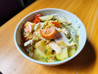 A bowl of Indonesian chicken soto with shredded chicken, sliced boiled egg, vermicelli, sliced cabbage, bean sprouts, celery, chili sauce and lime, mixed with delicious sauce. On a wooden table.	
