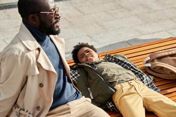 Portrait of handsome African American father and son spending time together outdoors relaxing on bench enjoying sunny weather