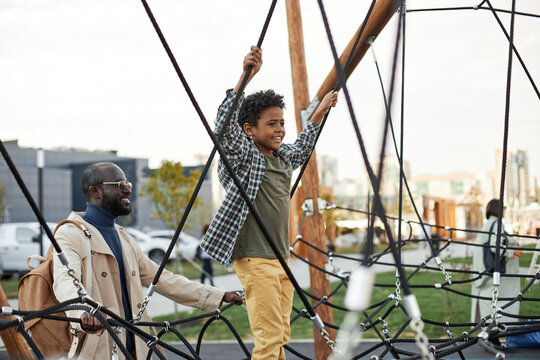 Horizontal shot of modern African American man spending free time with his son outdoors having fun on playground
