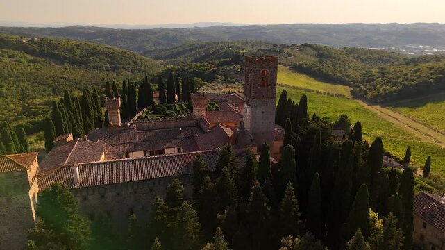 Drone sunset view of medieval Tuscan villa with bell clock tower, terracotta roofs, cypress avenue, formal Italian garden landscape design, surrounded by graphic green plowed fields in Italy