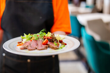Waiter in orange shirt and black apron holding plate with sliced roast beef, cherry tomatoes, and greens in restaurant setting with teal chairs