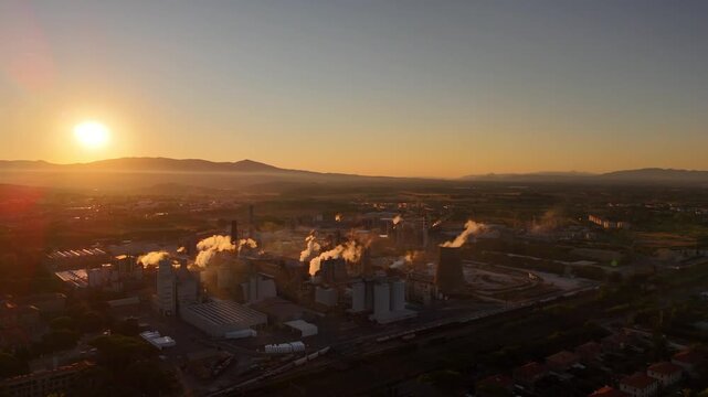 Drone aerial view of massive industrial factory with smokestacks, sprawling grounds, machinery and production lines at sunset, emphasizing industrial scale and power