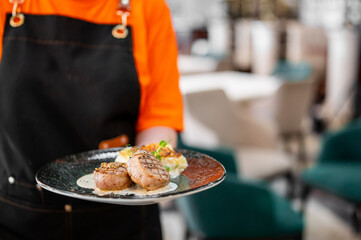 Waiter in orange shirt and black apron holding a plate with grilled steak medallions, creamy sauce, and mashed potatoes in a restaurant setting.