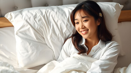 Beautiful young pretty Asian woman wake up and make happy smile with white shirt at the white bed in the morning.