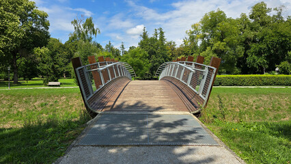 Modern wood and steel footbridge with gently sloping ramp connects park trails, framed by fresh green grass and mixed tree species in a well-kept city park