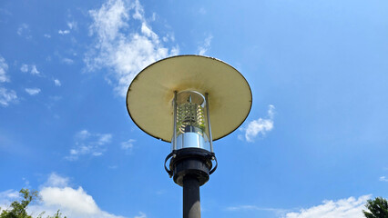 Stylish circular lamp head with metallic shaft stands out against bright blue sky and white clouds, designed for efficient lighting in urban park spaces