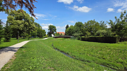 A gentle stream with a small footbridge and winding park paths is framed by lush green lawns, mature trees, and blue sky, inviting a scenic stroll in summer