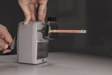 One hand is sharpening a wooden pencil using a grey pencil sharpener.