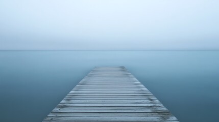 Fototapeta premium A long wooden pier extends into a calm, misty lake under a pale sky.