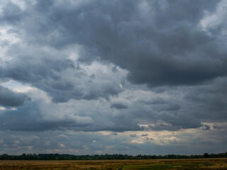 A thin band of marshland horizon lays across the lower image below a large area of dark cumulonimbus storm clouds  with sunlight breaking through in places.