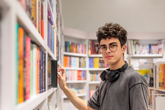 Teenage student with glasses and headphones browsing books in bookstore or library. education, research, academic lifestyle, self development, reading and knowledge